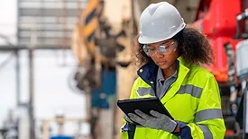 This image shows a person in personal protective equipment (PPE) using a tablet, likely in an industrial or construction setting