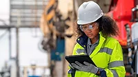 This image shows a person in personal protective equipment (PPE) using a tablet, likely in an industrial or construction setting