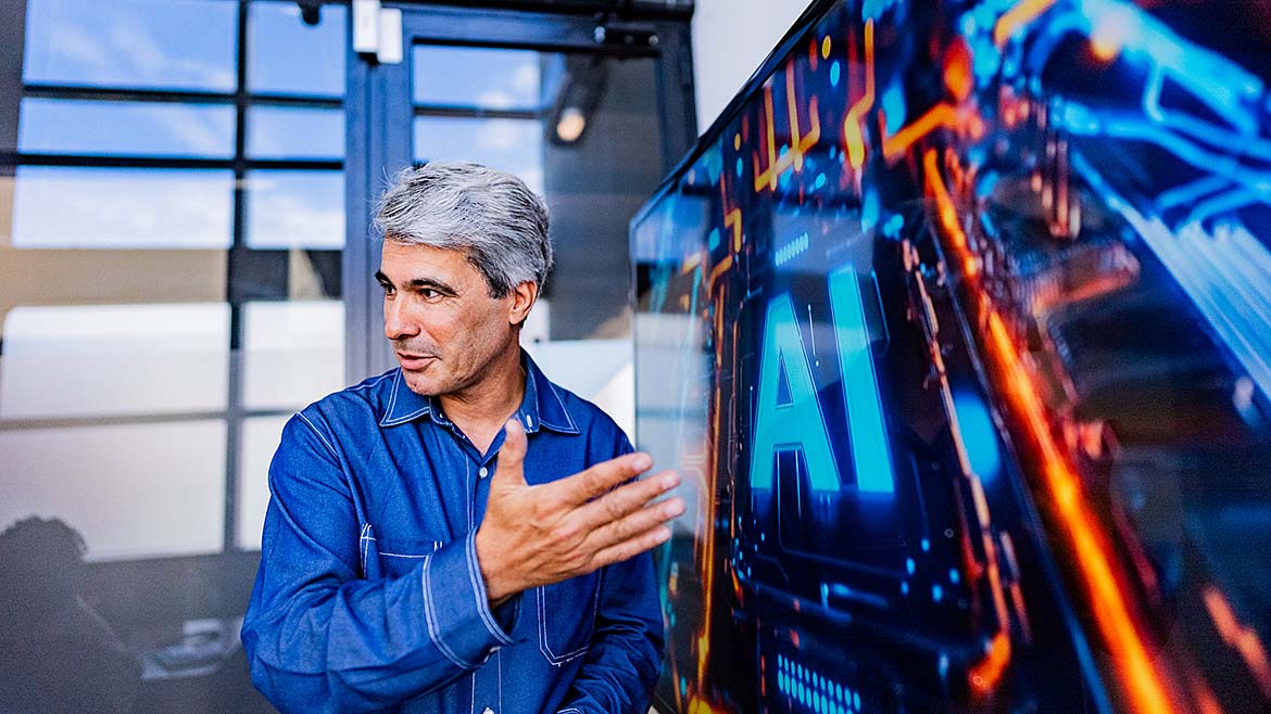The image depicts a man speaking in a modern setting with a large digital display featuring the letters 