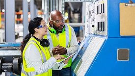 The image shows two multi-ethnic workers in a factory setting at a control panel. 