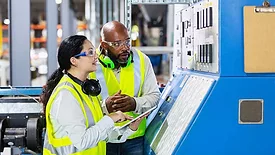 The image shows two multi-ethnic workers in a factory setting at a control panel. 
