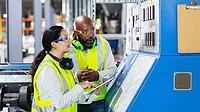 The image shows two multi-ethnic workers in a factory setting at a control panel. 