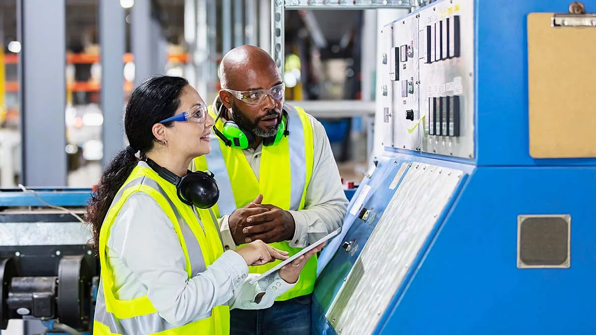 The image shows two multi-ethnic workers in a factory setting at a control panel. 