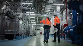 This image shows two engineers in high-visibility safety vests and hard hats inspecting industrial machinery. 