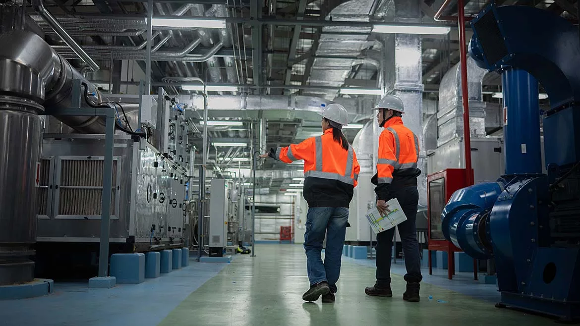 This image shows two engineers in high-visibility safety vests and hard hats inspecting industrial machinery. 