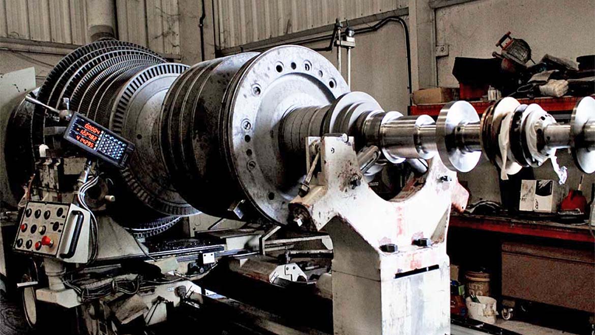 The image shows a large steam turbine rotor undergoing maintenance in a workshop. 