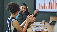 This image depicts a professional business meeting where a diverse group of people are applauding, likely celebrating a positive result or achievement. 