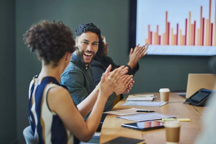 This image depicts a professional business meeting where a diverse group of people are applauding, likely celebrating a positive result or achievement. 