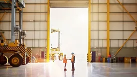 The image is a stock photo of a business interaction in an industrial warehouse setting. 