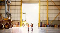 The image is a stock photo of a business interaction in an industrial warehouse setting. 