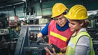 This image shows two industrial workers in a manufacturing facility reviewing information on a tablet computer. This image shows two industrial workers in a manufacturing facility reviewing information on a tablet computer.