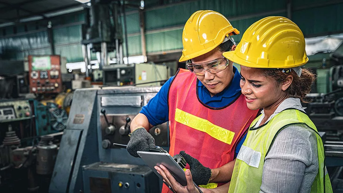 This image shows two industrial workers in a manufacturing facility reviewing information on a tablet computer.