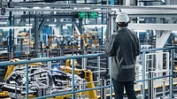 This image depicts a car factory engineer wearing a work uniform and a hard hat while using a tablet computer to oversee vehicle production with robotic arms. This image depicts a car factory engineer wearing a work uniform and a hard hat while using a tablet computer to oversee vehicle production with robotic arms.