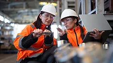 This image shows two individuals in high-visibility orange jackets and white hard hats collaborating in an industrial setting.
