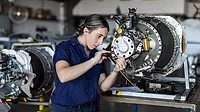 This image depicts a female aircraft engineer performing maintenance on a complex jet engine component in a hangar setting. 
