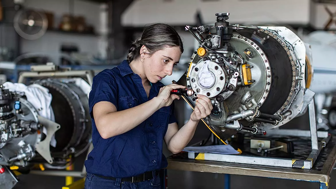 This image depicts a female aircraft engineer performing maintenance on a complex jet engine component in a hangar setting.