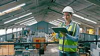 This image depicts a manufacturing engineer or worker using a tablet to update records within a factory setting. 