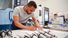 This image shows a person working with technical drawings and industrial machinery in a manufacturing setting. 