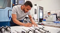 This image shows a person working with technical drawings and industrial machinery in a manufacturing setting. 