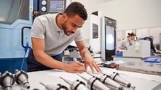 This image shows a person working with technical drawings and industrial machinery in a manufacturing setting. 