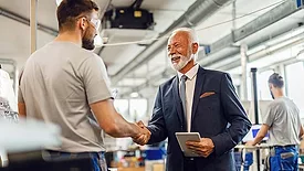 This image shows a senior businessman in a suit shaking hands with a factory worker in a manufacturing plant. 