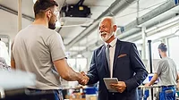 This image shows a senior businessman in a suit shaking hands with a factory worker in a manufacturing plant. This image shows a senior businessman in a suit shaking hands with a factory worker in a manufacturing plant.
