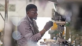 The image depicts a worker operating a punching or stamping machine within a heavy metal industry factory. 