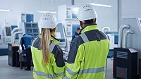 industrial professionals in safety gear discussing a process on a factory floor.