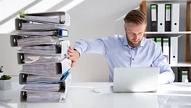 a person pushing away a large stack of paper files while working on a laptop
