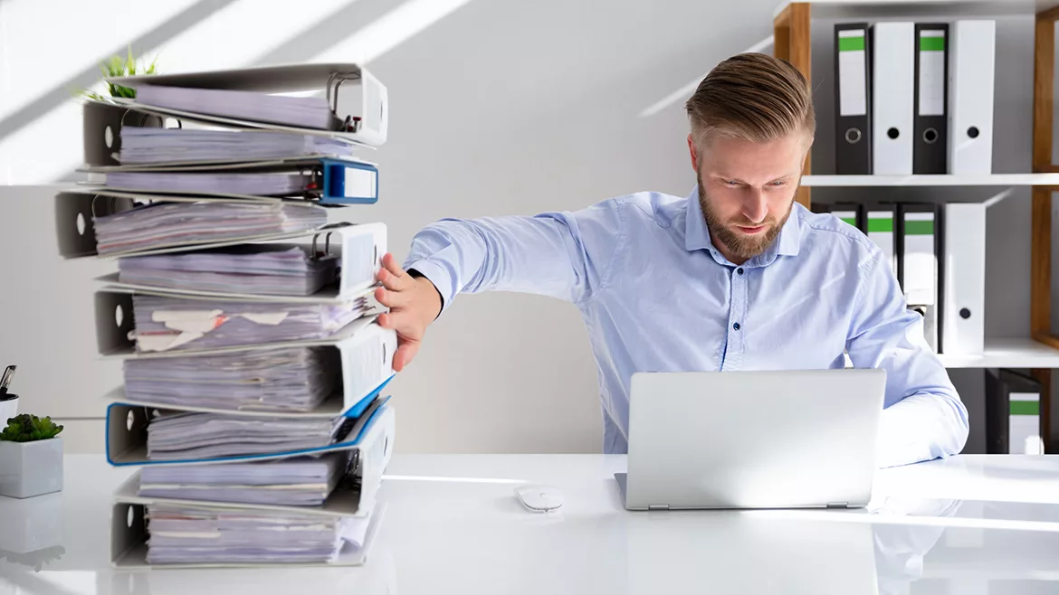 a person pushing away a large stack of paper files while working on a laptop