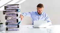 a person using a laptop while pushing away a large stack of office files